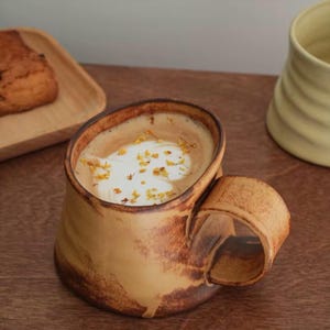 Puede incluir: Una taza de cerámica marrón hecha a mano con un asa única, llena de una bebida cremosa y cubierta con pequeños pétalos de flores amarillas. La taza está sobre una superficie de madera, con una bandeja de madera y otra taza de cerámica en el fondo.