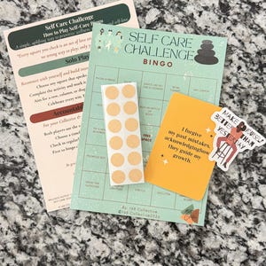 May include: A Self Care Challenge Bingo game set including an instruction sheet, a green bingo card, a strip of ten circular beige stickers, a yellow affirmation card with text, and a die-cut sticker featuring a person and the text Make Yourself Better Than Yesterday.