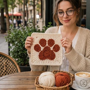 May include: A square crochet motif featuring a rust-colored paw print design centered on a cream-colored background. The piece is crafted with textured yarn stitches, creating a distinct contrast between the dark paw shape and the light border.