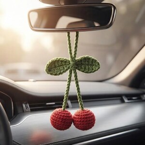 May include: A crochet cherry car hanging featuring two red spherical cherries attached to green stems and two green leaves. The text CROCHET CHERRY CAR HANGING and PDF PATTERN is visible. The item hangs from a rearview mirror inside a vehicle.