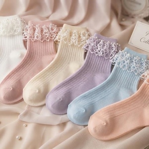 Könnte beinhalten: Ein Set aus sechs gerippten Socken in Weiß, Rosa, Creme, Lavendel, Hellblau und Pfirsich. Jede Socke ist am Bündchen mit einer dekorativen weißen Spitzenrüsche versehen. Eine kleine weiße Karte trägt die Aufschrift Pastel Collection.