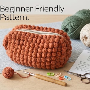 May include: A rust-colored crocheted pouch with a bobble stitch texture and a zipper closure. The text "Beginner Friendly Pattern." is visible. Included are a wooden crochet hook, four plastic stitch markers, scissors, and yarn balls.
