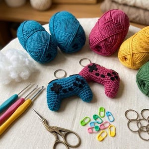 May include: A selection of crochet items including balls of blue, pink, yellow, and green yarn, crochet hooks, scissors, stitch markers, and keyrings shaped like game controllers. The keyrings are blue and pink.