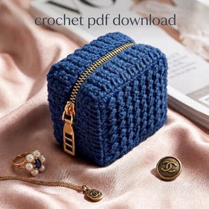 May include: A small, rectangular, dark blue crochet pouch featuring a gold-toned metal zip closure. The text 'crochet pdf download' is visible at the top. A gold ring with white pearls and a blue stone, and a gold button are nearby.