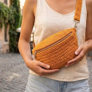 Può includere: Marsupio all'uncinetto in una calda tonalità arancione, con cerniera diagonale e cinturino regolabile. La borsa è tenuta da una persona, mostrando il suo design strutturato e lo stile casual. Le dimensioni della borsa non sono visibili.