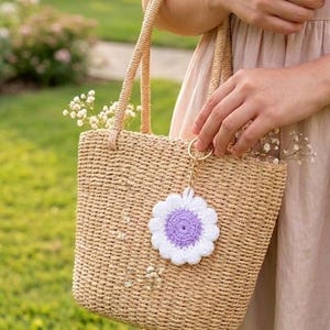 May include: A woven straw tote bag with a crochet flower charm. The bag has a rectangular shape with a woven handle. The flower charm is white with a purple center. The text "crochet pdf download" is at the bottom.