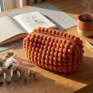 May include: A rectangular, burnt orange zippered pouch featuring a textured, bobble-stitch crochet pattern. The pouch has a structured, boxy shape with a visible metal zipper closure running along the top edge, showcasing a dense, uniform knit design.