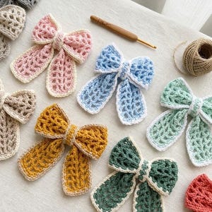 Puede incluir: Una colección de lazos de ganchillo hechos a mano en varios colores, incluyendo rosa, azul, amarillo y verde. Cada lazo presenta un delicado diseño de tejido abierto con un ribete blanco contrastante. También se ven un ganchillo y cuerda.