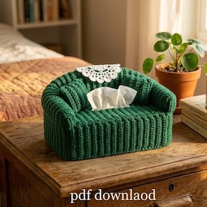 May include: A green, crocheted tissue box cover shaped like a miniature sofa, with white tissues and a white lace doily. The tissue box is on a wooden table, with a plant and a mug that says "Tea" in the background.