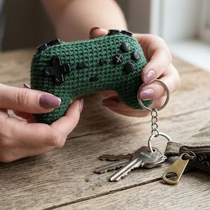 Könnte beinhalten: Ein handgefertigter, grüner Häkel-Videospiel-Controller-Schlüsselanhänger mit schwarzen Knöpfen und einem silbernen Schlüsselring. Der Schlüsselanhänger wird in der Nähe eines Schlüsselsatzes und eines kleinen Canvas-Beutels auf einer Holzoberfläche gehalten.