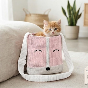 May include: A light pink and white crocheted bag with a fox face design, featuring black embroidered eyes and a black nose. A small orange kitten sits inside the bag. A tag in the corner reads: BESTSELLER DIGITAL CROCHET PATTERN PDF.