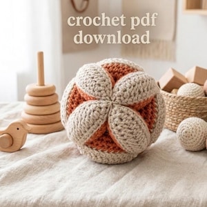 Op de afbeelding: Een handgemaakte gehaakte bal in beige en oranje, met de tekst "crochet pdf download". De afbeelding toont ook houten speelgoed, waaronder een stapelring en een vogel, op een neutrale ondergrond.