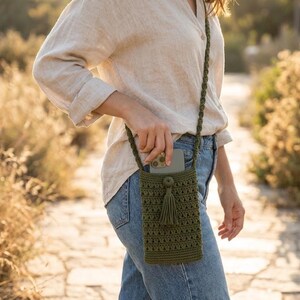 May include: Olive green crochet phone crossbody bag with a long strap and tassel. The bag is designed to hold a smartphone and is made of a textured, woven material. The bag is approximately 18 cm tall.