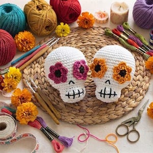 Könnte beinhalten: Zwei gehäkelte weiße Totenköpfe mit gestickten schwarzen Nähten für Nase und Mund. Ein Totenkopf hat pinkfarbene Blumen als Augenhöhlen, der andere orangefarbene. Sie sind umgeben von Garnknäueln, Häkelnadeln und orangefarbenen Ringelblumen.
