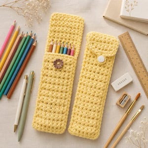 May include: Two yellow crochet pencil cases with a textured stitch pattern. One case is open, revealing six colored pencils, and features a brown button closure. The other case is closed with a white snap button. Both are rectangular and slim.