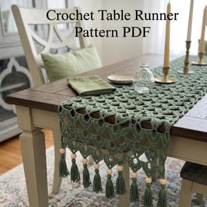 May include: A green crochet table runner with a scalloped edge and tassels, adorned with wooden beads. The text "Crochet Table Runner Pattern PDF" is displayed. The runner is placed on a wooden table with candles and a small glass bottle.
