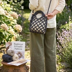 May include: A dark gray crocheted net bag with a shoulder strap, held by a person. The bag has a round handle and wooden beads. A book titled "Beginner Friendly Pattern" and yarn balls are on a terracotta pot.