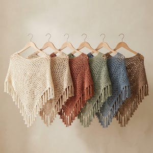 May include: Six crochet ponchos hanging on wooden hangers, each featuring an open-knit pattern and a pointed hem with fringe. The ponchos are displayed in a row, showcasing a color palette of cream, beige, terracotta, sage green, blue, and brown.