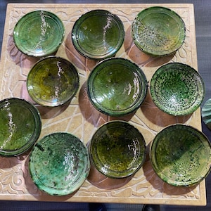 May include: Overhead shot of eleven green ceramic bowls with varying shades and textures. The bowls are arranged on a light-colored, patterned surface. The bowls have a glossy finish and appear to be handmade.