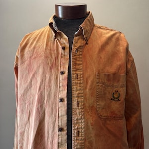 May include: An orange and pink button-down shirt with a chest pocket and embroidered emblem. The shirt has a classic collar, long sleeves, and brown buttons. The fabric appears to have a faded or distressed effect.
