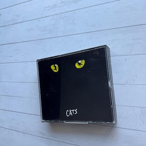 May include: A black CD case with the word "CATS" in white. The case has a black background with two yellow cat eyes. The case is on a light blue wooden surface.