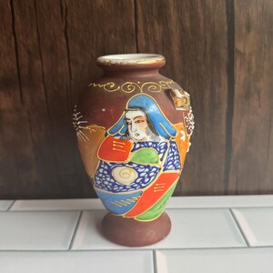 May include: A small brown ceramic vase featuring a hand-painted figure wearing a blue head covering and a colorful patterned robe. The vase has a narrow neck with a gold-painted rim and decorative gold accents around the upper body and base.
