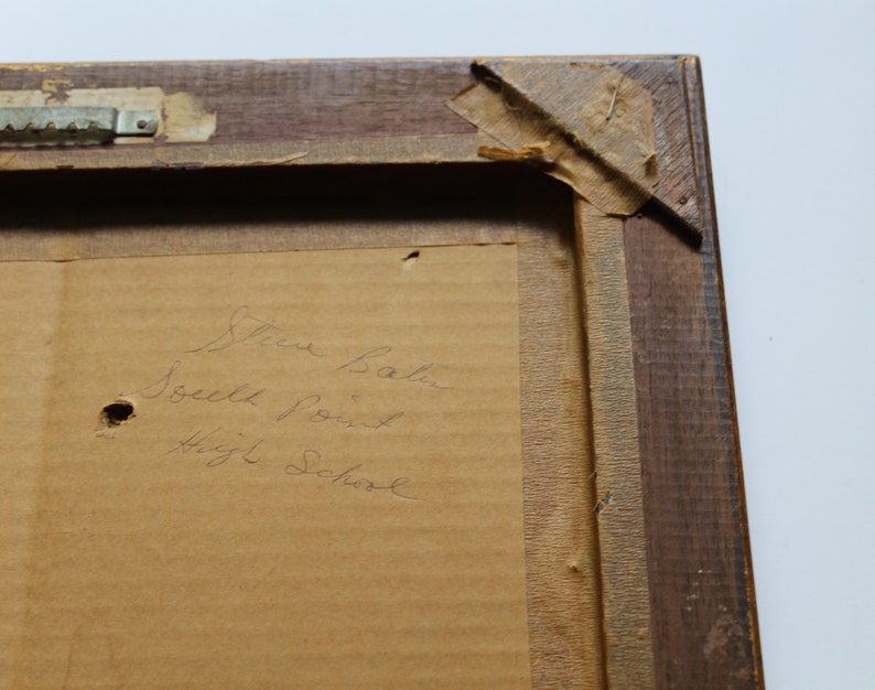 May include: A close-up of a wooden picture frame with a brown paper backing. The paper has handwritten text that reads "Steve Baker South Point High School".