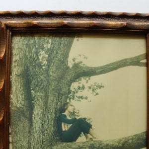 May include: A framed photograph of a woman sitting on a tree branch. The woman is wearing a dark blue shirt and black pants. The tree is large and has a thick trunk. The background is a light green.