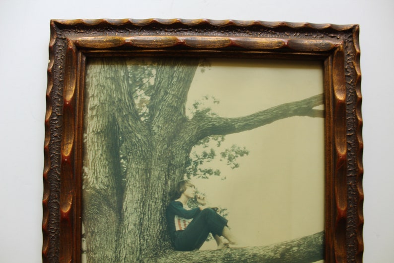 May include: A framed photograph of a woman sitting on a tree branch. The woman is wearing a dark blue shirt and black pants. The tree is large and has a thick trunk. The background is a light green.