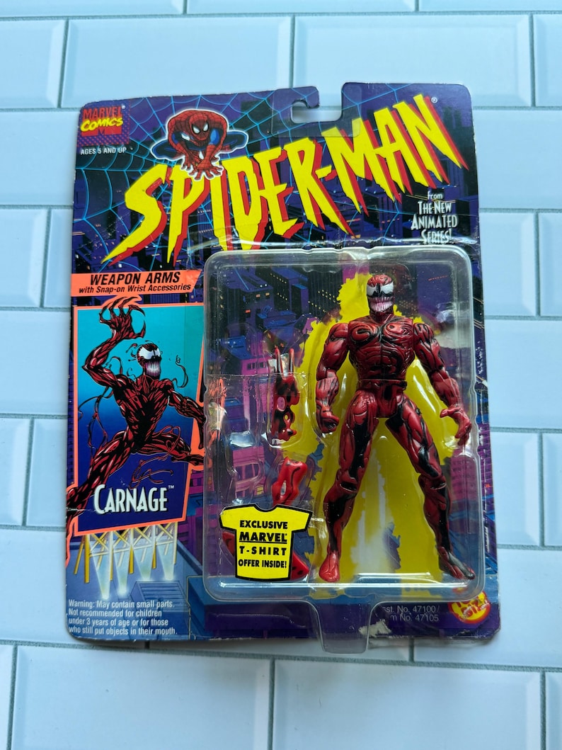 May include: A packaged Carnage action figure from the Spider-Man animated series. The red figure with black accents is displayed with weapon accessories. The packaging features the Spider-Man logo and text: "Exclusive Marvel T-Shirt Offer Inside!" The figure is in a plastic case.