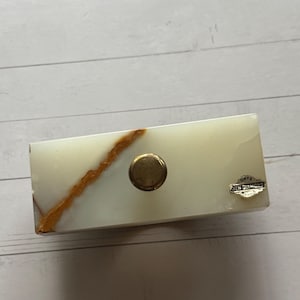 May include: Rectangular onyx box with a gold-colored knob. The box is off-white with a prominent brown vein running diagonally across the surface. A small silver tag reads "ONYX NEW DIAMOND".