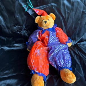 May include: A plush teddy bear dressed in a clown costume featuring a red and purple colour scheme with a checkered pattern and star accents. The bear wears a matching pointed hat with a teal ribbon and a ruffled collar around its neck.