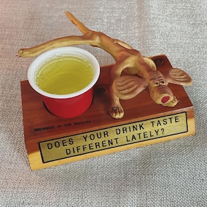 May include: A wooden shot glass holder featuring a cartoon dog figure. The holder is inscribed with the text "Does your drink taste different lately?" and "Cherokee in the Smokies".