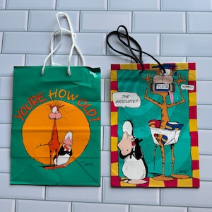May include: Two gift bags with cartoon illustrations. One is green with the text "YOU'RE HOW OLD?" and an orange circle with a cat and penguin. The other is red, yellow, and green with a cat and penguin and the text "THE GRADUATE?"