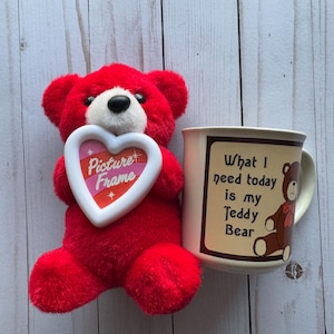 Puede incluir: Un oso de peluche rojo sosteniendo un marco de fotos blanco en forma de corazón con rayas rosas y naranjas y las palabras "Picture Frame" en su interior. El oso está sentado junto a una taza blanca con un borde y un asa marrones. La taza tiene un borde marrón alrededor del texto "What I need today is my Teddy Bear" y un oso de peluche marrón con una corbata de lazo roja.