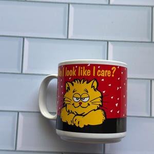 Puede incluir: Taza de cerámica blanca con una ilustración de gato amarillo y el texto "Do I look like I care?" sobre un fondo rojo con puntos blancos. La taza tiene una franja negra en la parte inferior y un asa blanca.