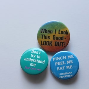 May include: Three round button pins with different sayings. The top pin is rainbow colored with the text "When I Look This Good - LOOK OUT!" The left pin is teal blue with the text "Don't try to understand me Just Love me". The right pin is blue with the text "PINCH ME PEEL ME EAT ME LOUISIANA CRAWFISH".