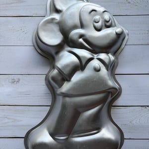 Könnte beinhalten: Eine silberne Mickey-Mouse-Kuchenform aus Metall. Die Form ist wie Mickey Mouse geformt, mit seinen charakteristischen Ohren, großen Augen und einem freundlichen Lächeln.