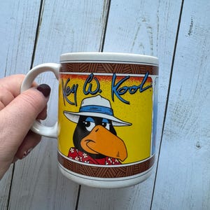 May include: White ceramic mug featuring a cartoon illustration of a black bird wearing a white and blue hat and a red and white Hawaiian shirt. The text "Key West Kool" is written in black on a yellow background.
