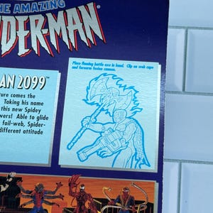 May include: A close-up of a blue and silver action figure package featuring "THE AMAZING SPIDER-MAN" logo. The package includes illustrations of Spider-Man characters and text that reads "AN 2099". A diagram shows how to place a flaming battle axe in hand.