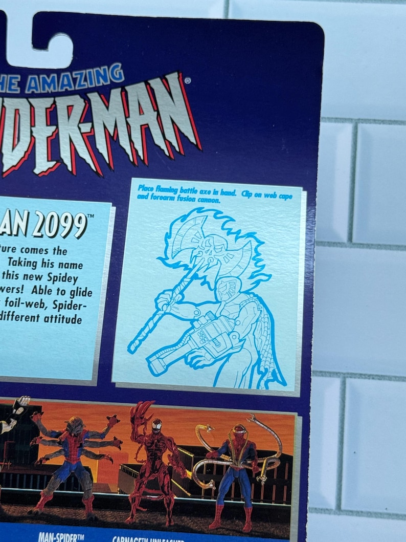 May include: A close-up of a blue and silver action figure package featuring "THE AMAZING SPIDER-MAN" logo. The package includes illustrations of Spider-Man characters and text that reads "AN 2099". A diagram shows how to place a flaming battle axe in hand.