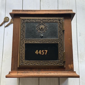 Pode incluir: Uma caixa de correio de madeira vintage com uma porta de metal escuro com o número "4457" em dourado. A porta tem uma borda ornamentada e geométrica e um emblema em forma de estrela. Duas chaves estão ao lado da caixa de correio.