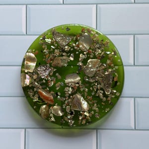 Puede incluir: Un posavasos redondo de resina verde con fragmentos de concha iridiscente incrustados y pequeñas piedras de colores. El posavasos tiene un acabado brillante. El diseño es decorativo y único, sobre un fondo de baldosas blancas.