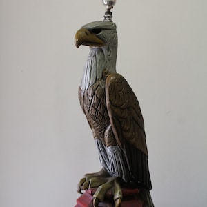 May include: A ceramic eagle lamp with a brown and grey painted design. The eagle is perched on a red base with a light bulb on top.