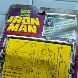 May include: Marvel Comics Iron Man action figure packaging featuring Hawkeye. The packaging is purple and yellow with illustrations of Hawkeye and his equipment. Text includes "Lord arrow in bow as shown. Pull back and release to fire."