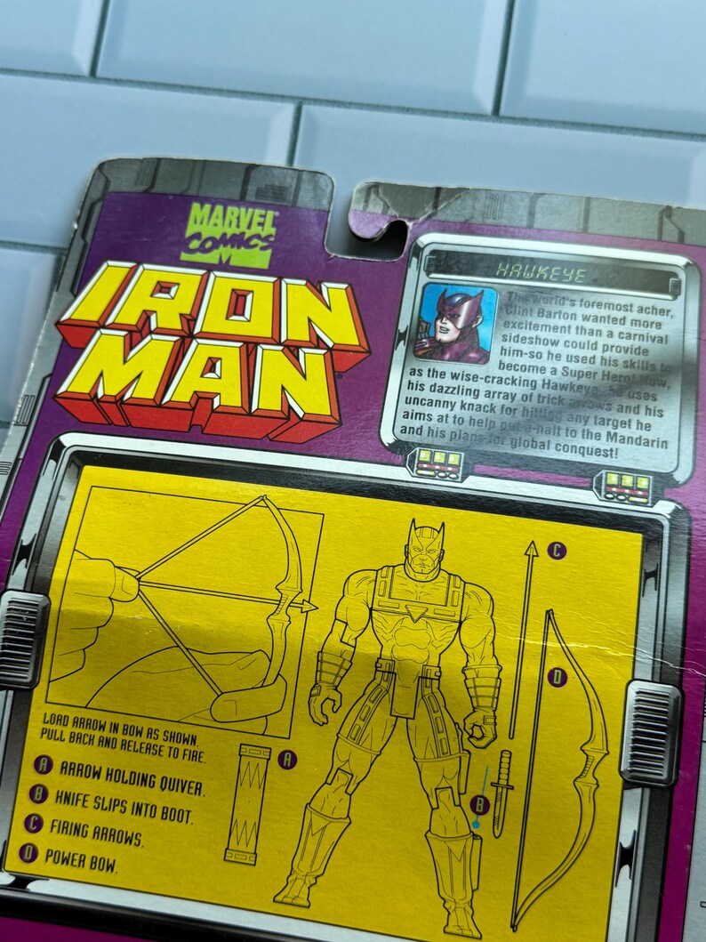 May include: Marvel Comics Iron Man action figure packaging featuring Hawkeye. The packaging is purple and yellow with illustrations of Hawkeye and his equipment. Text includes "Lord arrow in bow as shown. Pull back and release to fire."