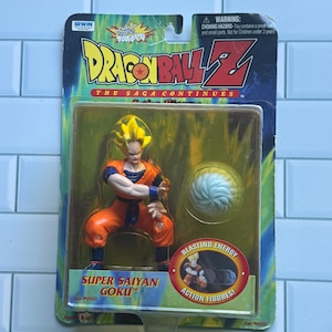 May include: A Dragon Ball Z Super Saiyan Goku action figure in original packaging. The figure has spiky yellow hair and an orange gi. Included is a white energy blast accessory. The card features the text Dragon Ball Z The Saga Continues.