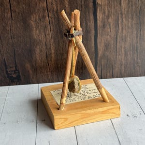 May include: A small wooden sculpture featuring three wooden sticks bound together, supporting a small stone. The sticks are attached to a wooden base with handwritten text. The sculpture has a rustic, natural aesthetic.