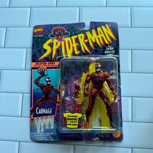 May include: A red and black Carnage action figure from the Spider-Man animated series, in its original packaging. The card features the Spider-Man logo and the text "Carnage". The packaging also includes an offer for a Marvel t-shirt.