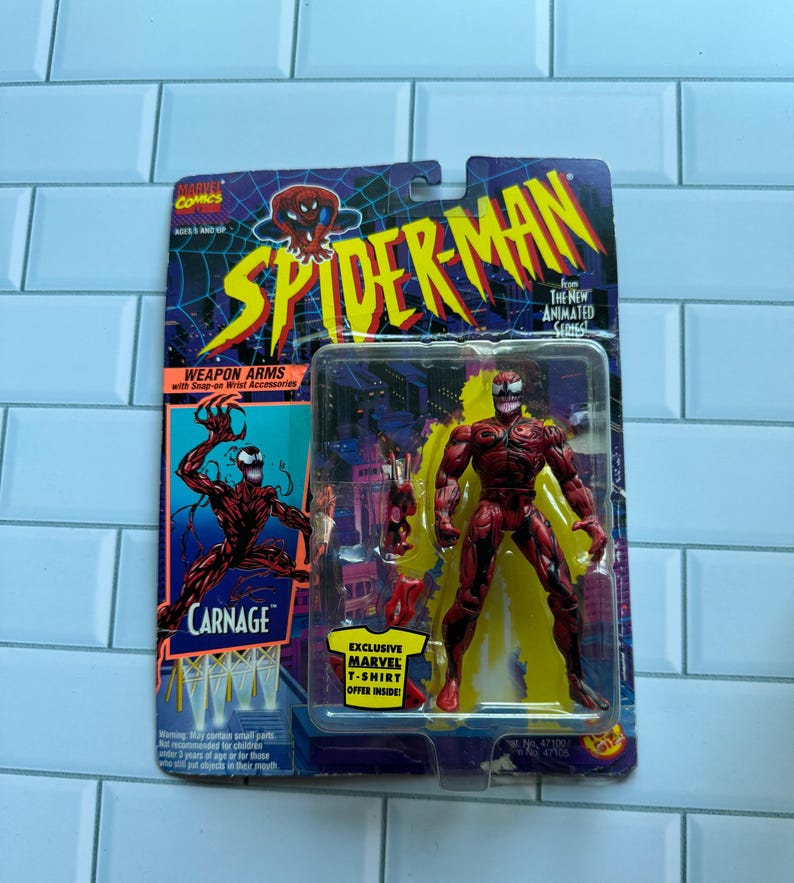 May include: A red and black Carnage action figure from the Spider-Man animated series, in its original packaging. The card features the Spider-Man logo and the text "Carnage". The packaging also includes an offer for a Marvel t-shirt.